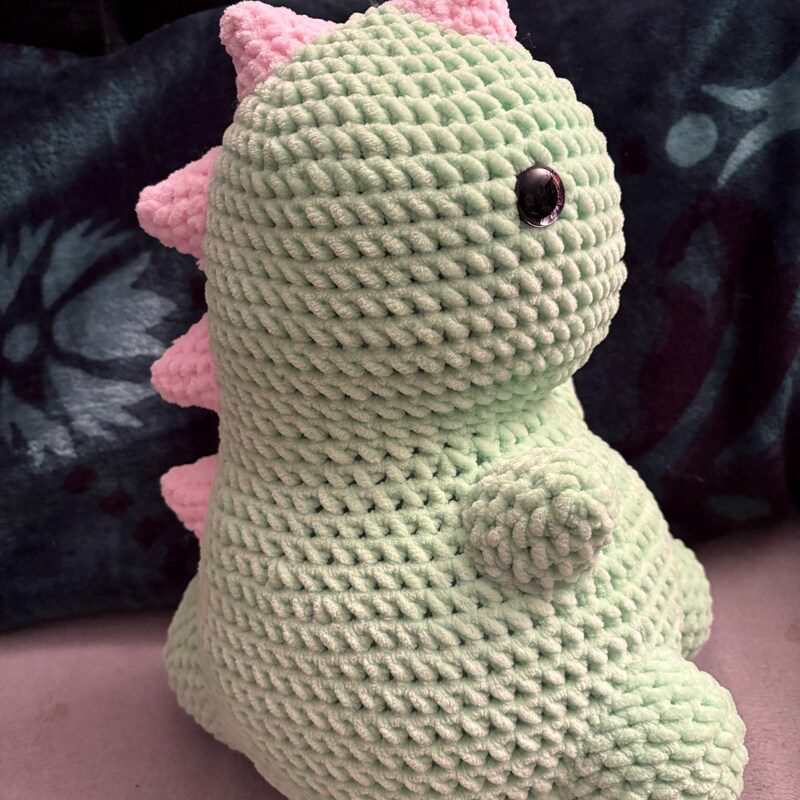 Chubby pink dinosaur amigurumi crochet patterns with a rounded body and small spikes, made with pastel yarn and clean crochet stitches.