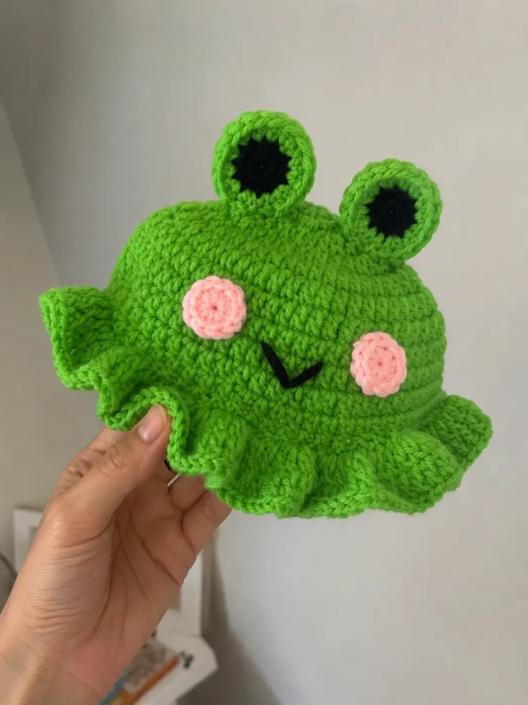 Finished crochet frog hat for cat pattern held in hand, showing the eyes, cheeks, and smile detail up close