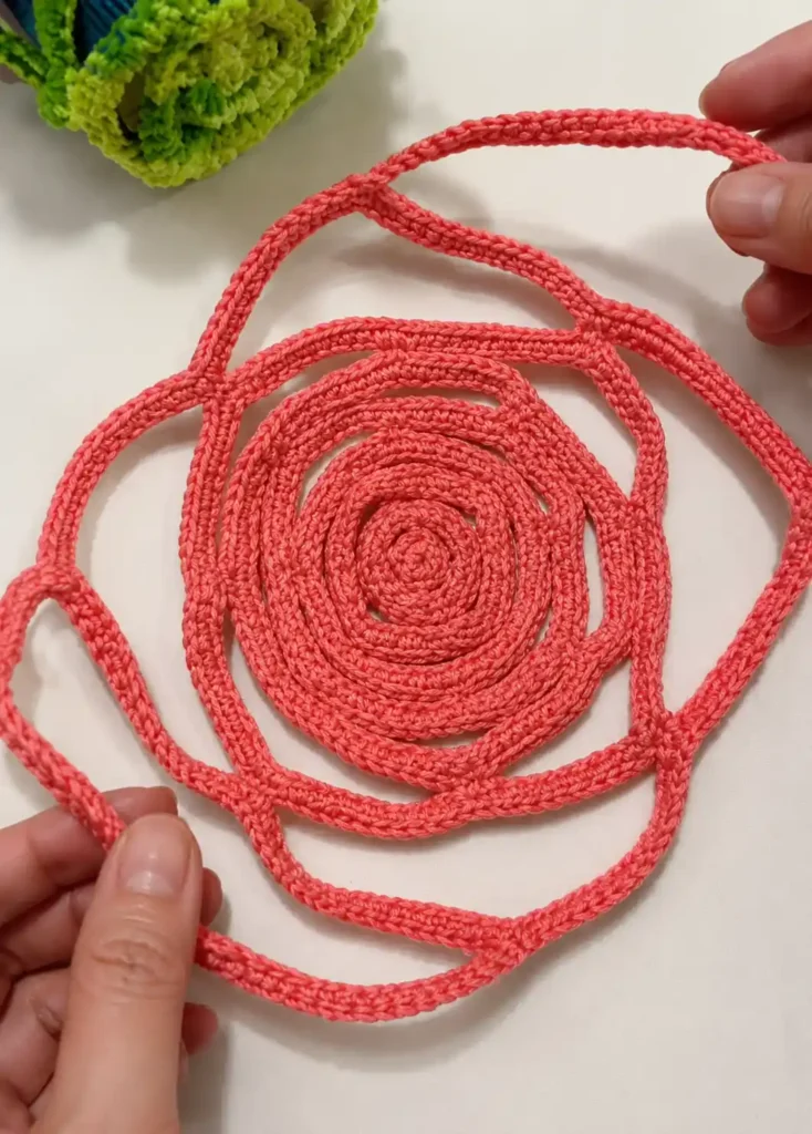 Crochet rose mesh cup holder pattern laid flat on a table in coral yarn, showing the spiral rose shape before the petals are pulled up into a mesh
